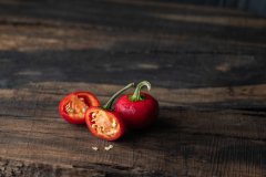 Red cherry pepper sliced in half showing seeds, beside a whole pepper on dark rustic wood