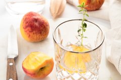 Fresh peach slices, whole peaches, and thyme around a crystal glass on a light marble counter