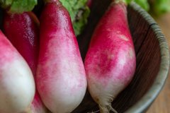 A bunch of fresh French breakfast radishes with pink and white skins and green tops in a dark wooden bowl