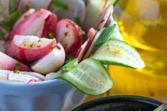 A radish and cucumber salad in a blue bowl tossed with grainy mustard vinaigrette, with a spoon of dressing and a bottle of olive oil beside it