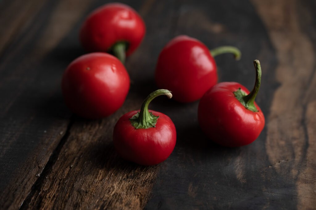 Five whole red cherry peppers with green stems on a dark rustic wooden table