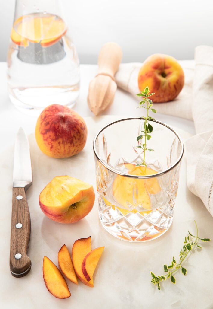 Fresh peach slices, whole peaches, and thyme around a crystal glass on a light marble counter