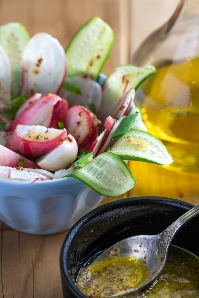 Radish and cucumber salad in a blue bowl tossed with grainy mustard vinaigrette, a spoon of dressing and a bottle of olive oil beside it