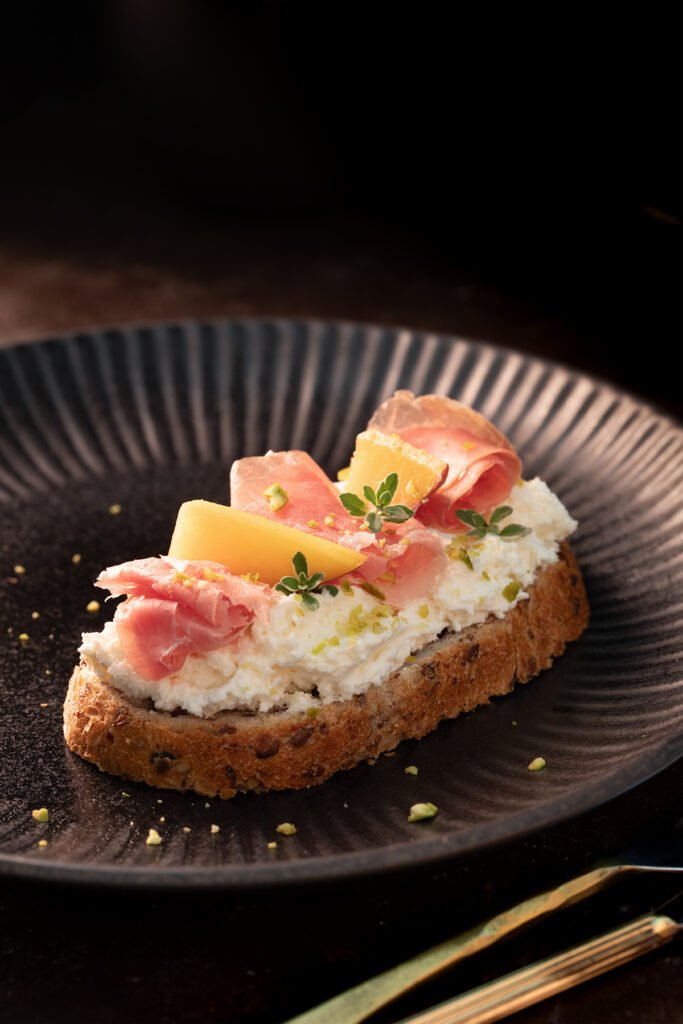 Creamy ricotta toast topped with prosciutto, fresh peach slices, thyme, and crushed pistachios on rustic seeded bread served on a black ceramic plate.