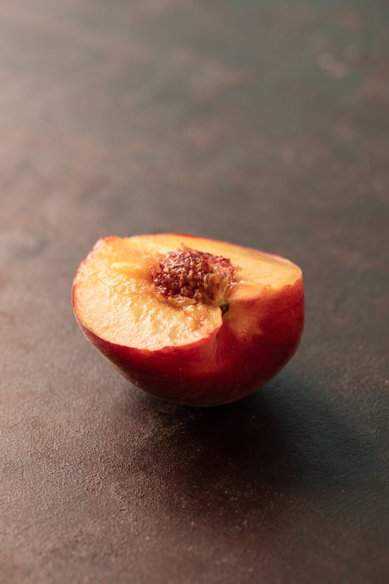 Ripe summer peach cut in half, showing juicy amber flesh and red-fibred stone, on a dark textured surface in soft directional light.