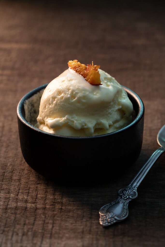 Scoop of peach ice cream in a black bowl topped with caramelised peach, vintage spoon alongside, on a rustic wooden table in warm directional light.