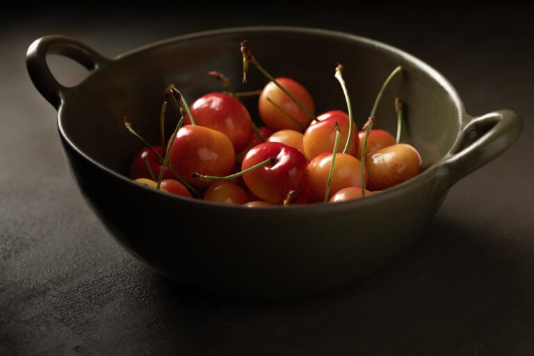 Rainier cherries with green stems in a dark green stoneware bowl on a black textured surface, moody chiaroscuro food photography