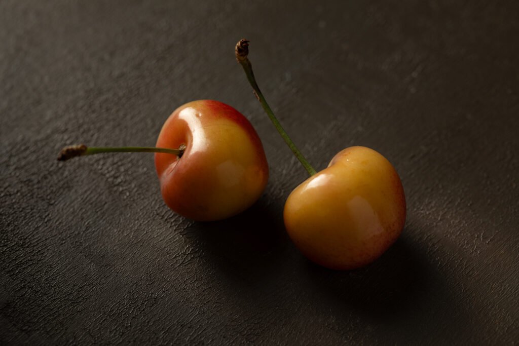 Two Rainier cherries with crossed green stems on a dark textured surface, minimalist chiaroscuro still life