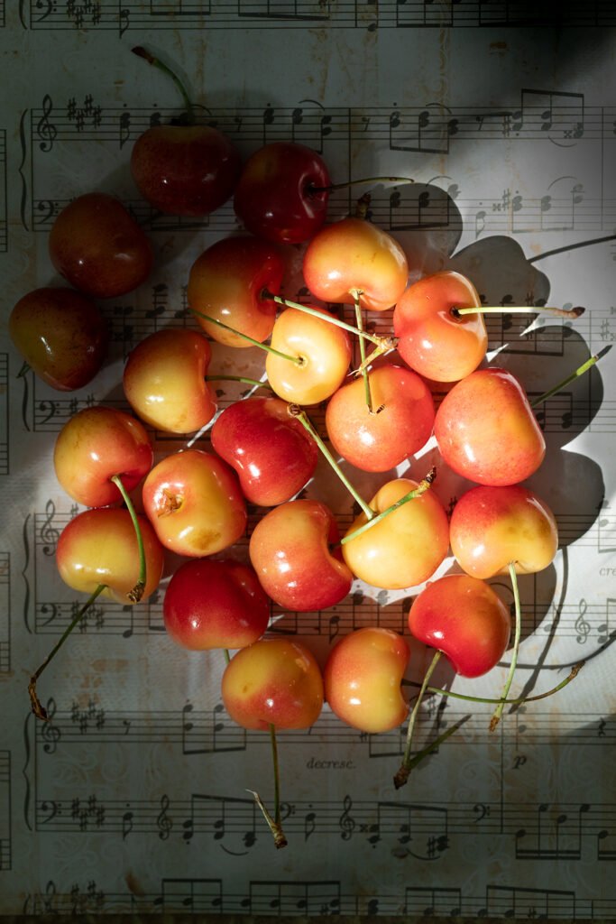 Rainier cherries scattered on vintage sheet music in hard directional sunlight with deep shadows, overhead flat lay still life