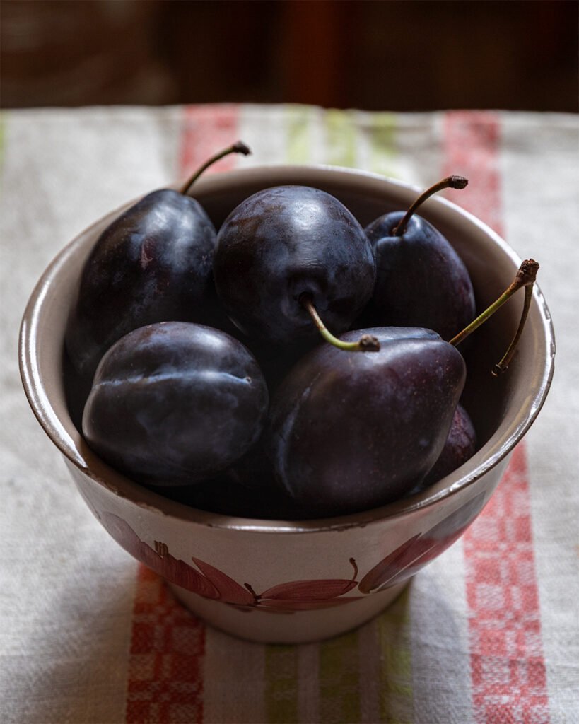 Dark purple plums with stems in a hand-painted ceramic bowl on a striped linen kitchen towel