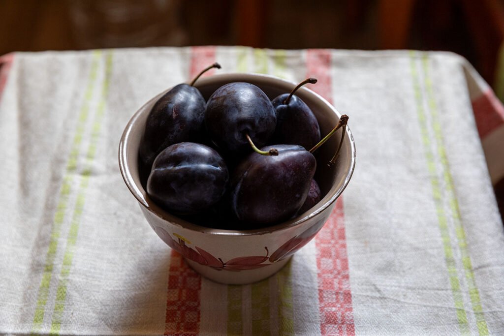 Before editing: dark plums in a hand-painted ceramic bowl on a bright striped linen kitchen towel